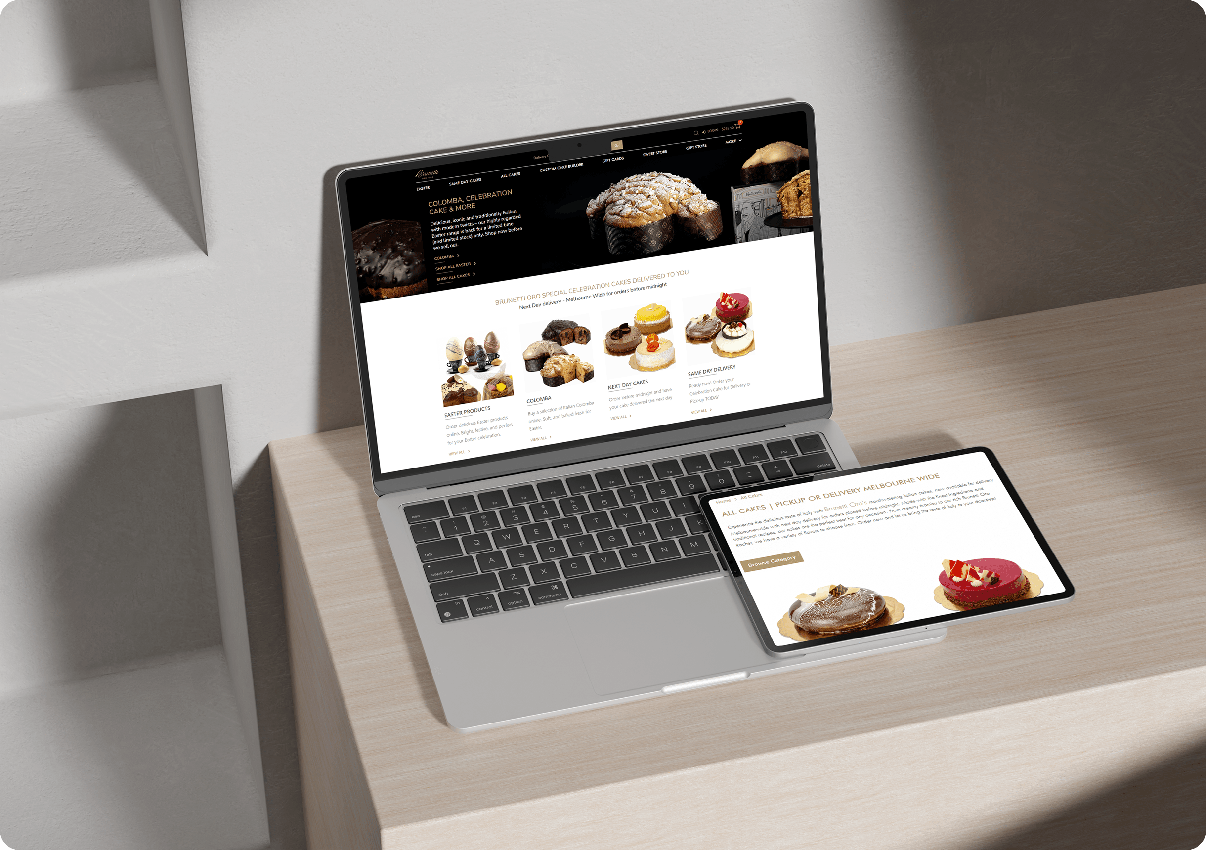 A laptop and a tablet on a light wood desk display a bakery website with images of pastries, breads, and desserts.