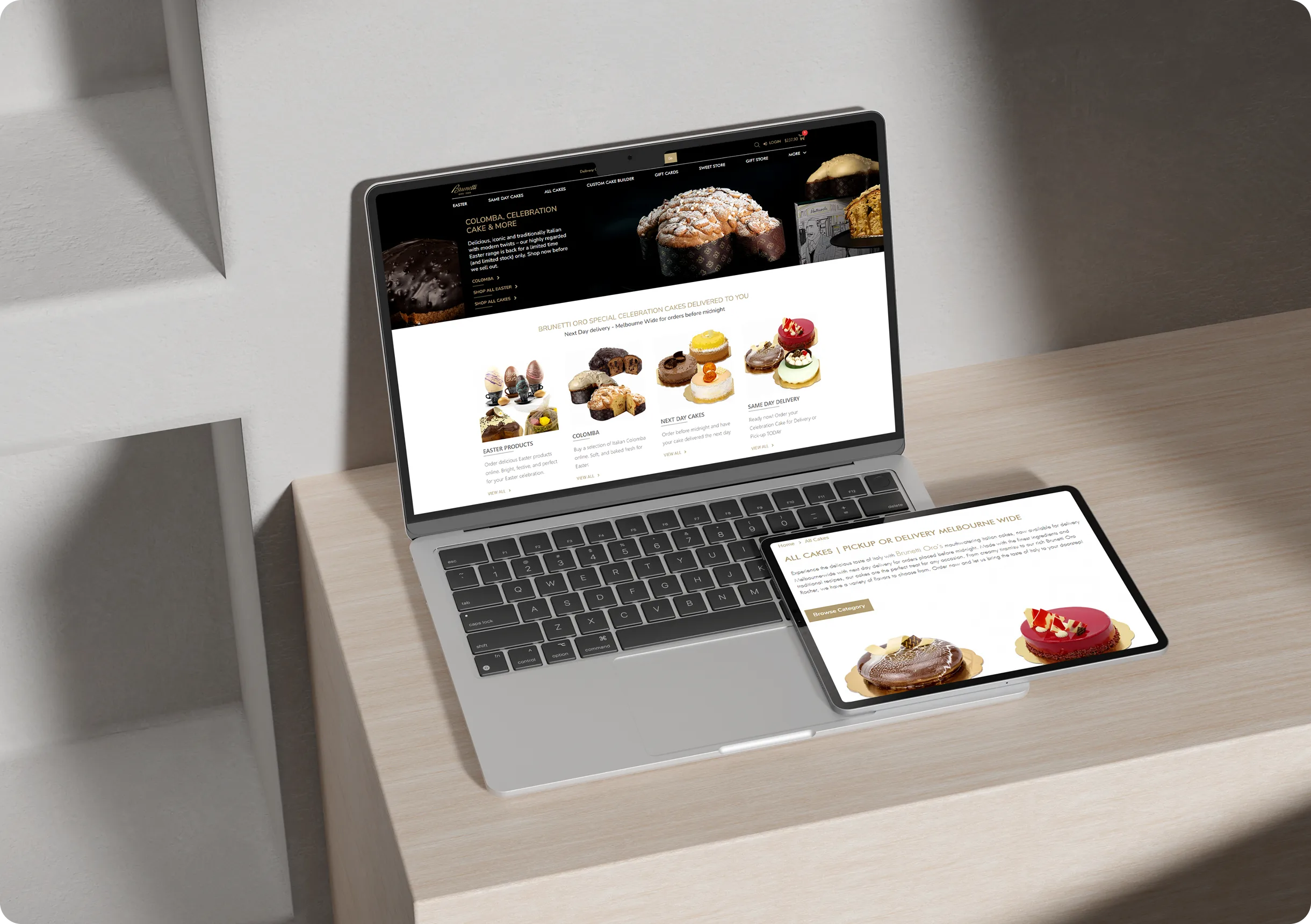 A laptop and a tablet on a light wood desk display a bakery website with images of pastries, breads, and desserts.