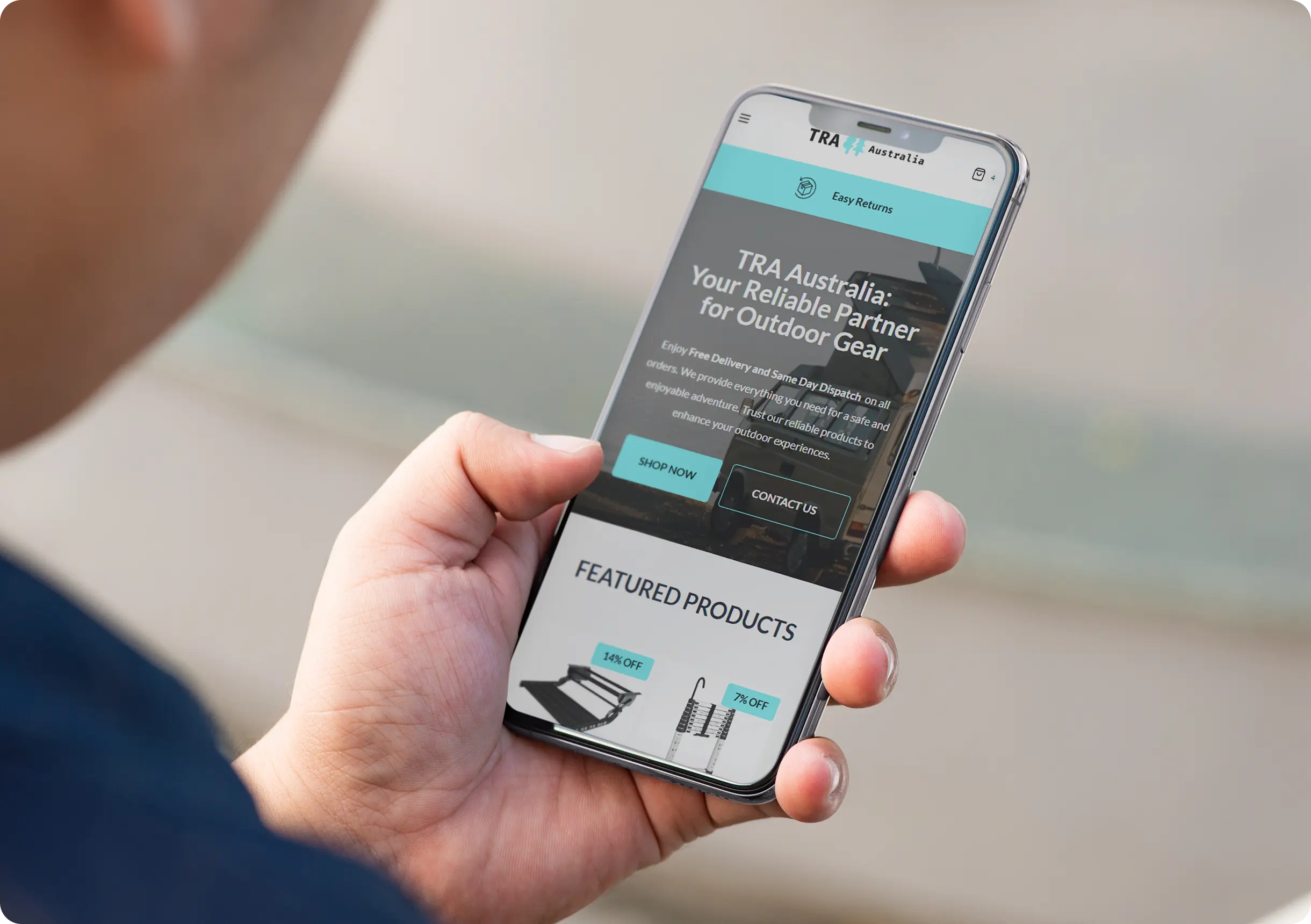 A person holds a smartphone displaying the homepage of TRA Australia’s website, which offers outdoor gear and features buttons for shopping and contacting.